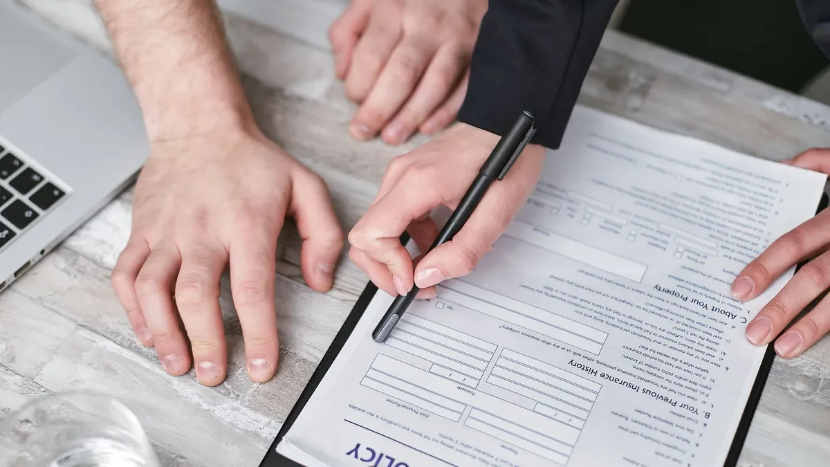 Hands completing an insurance policy form on a clipboard beside a laptop