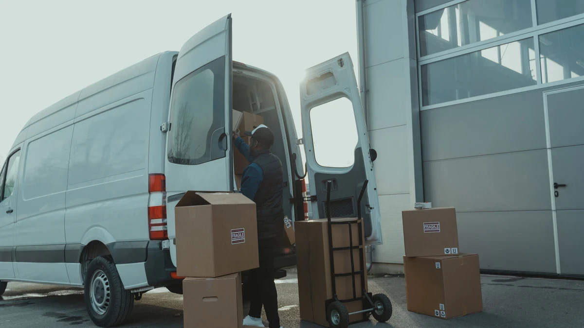 Courier loading cardboard boxes into the back of a white panel van outside a warehouse, a sack truck stacked beside him