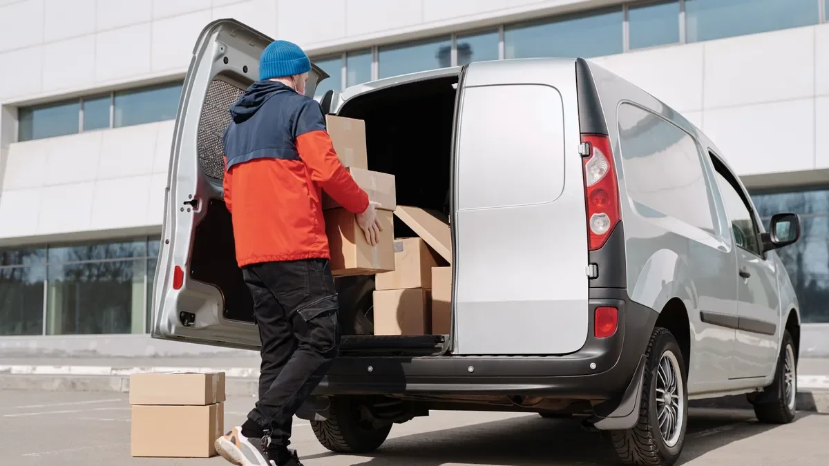 Courier in an orange jacket loading cardboard boxes into the back of a silver van