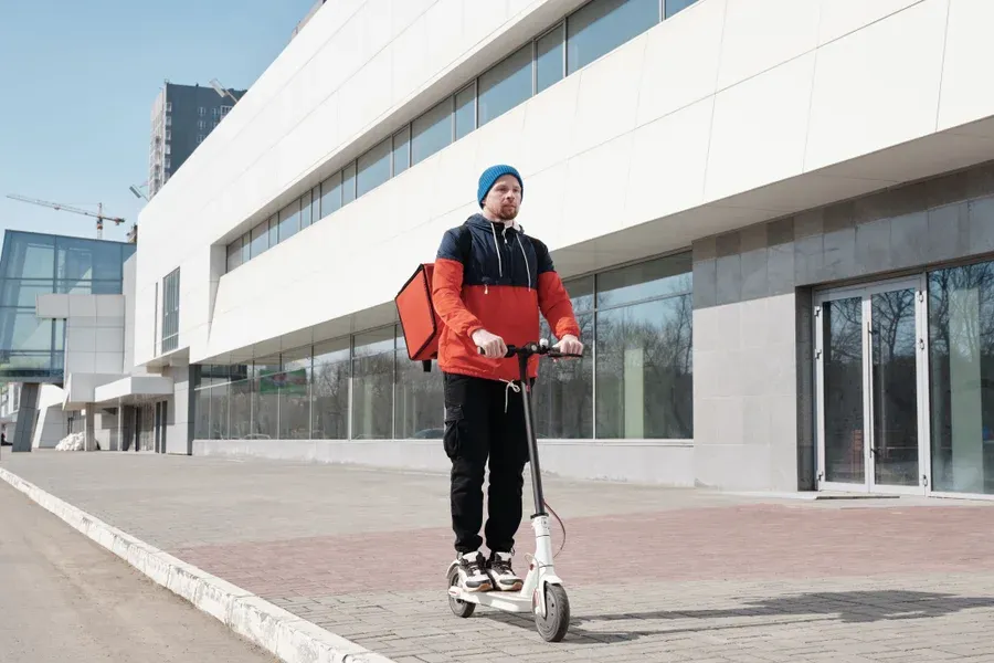 Courier riding an electric scooter with a red thermal delivery bag past a modern building