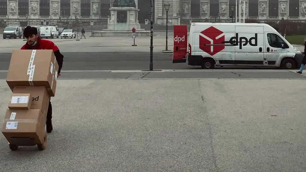 Courier wheeling a stack of parcels on a hand truck across a square with a DPD van parked behind