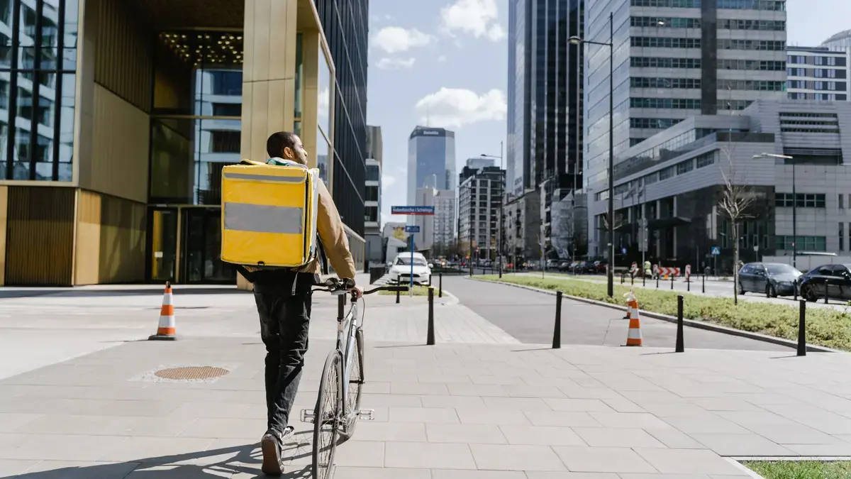 Cycle courier with a yellow thermal delivery backpack pushing his bike across a city plaza