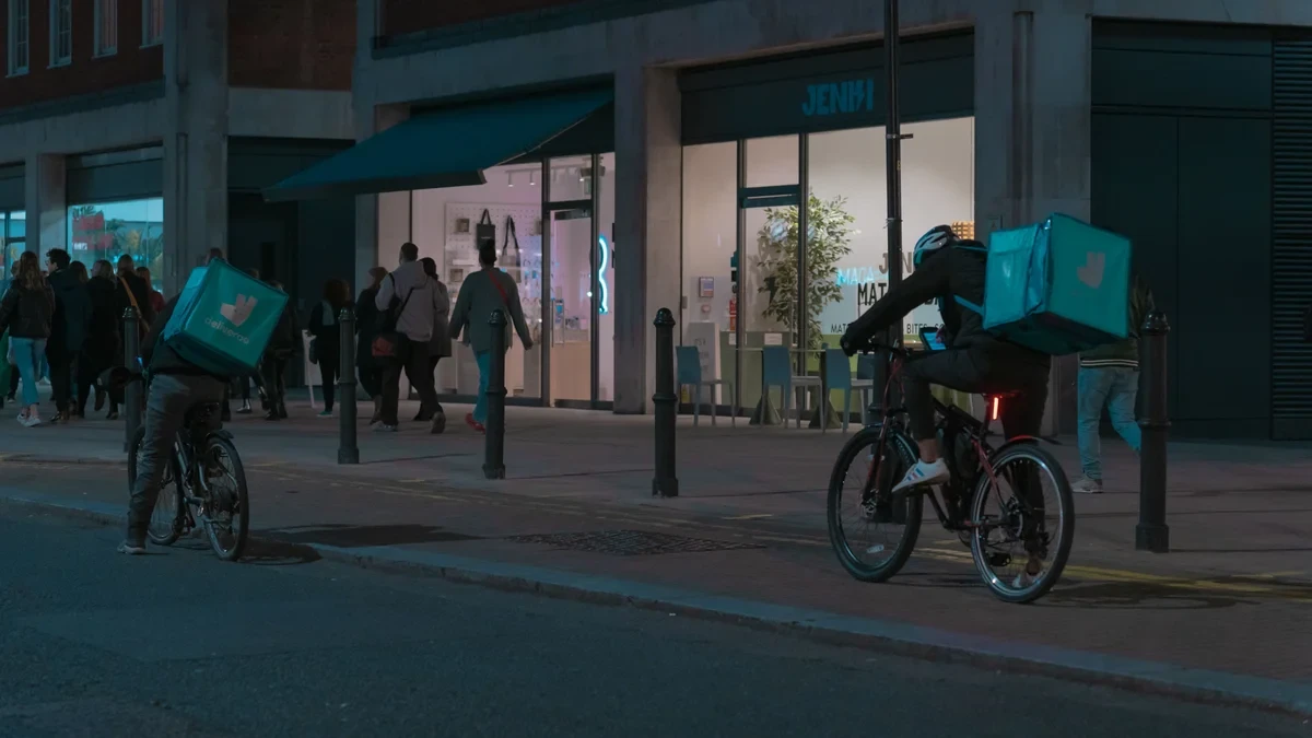 Two Deliveroo cyclists riding past lit shopfronts on a UK high street at night
