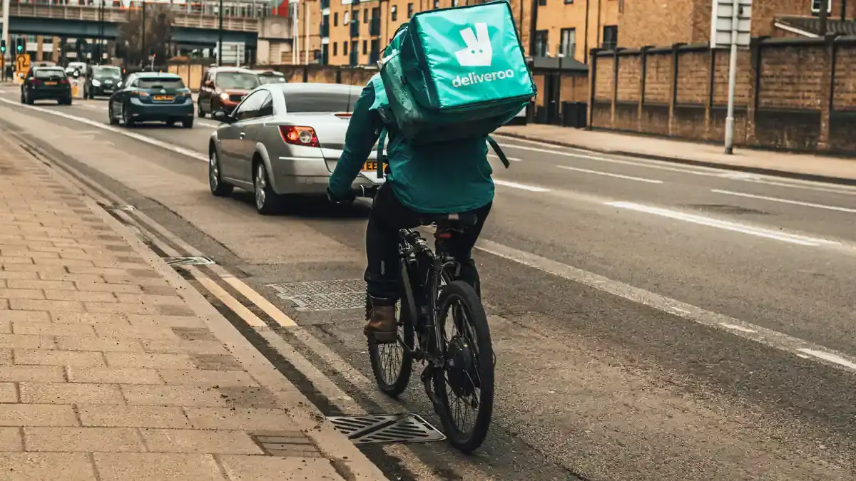 Deliveroo rider with a teal delivery box cycling along a UK street past traffic