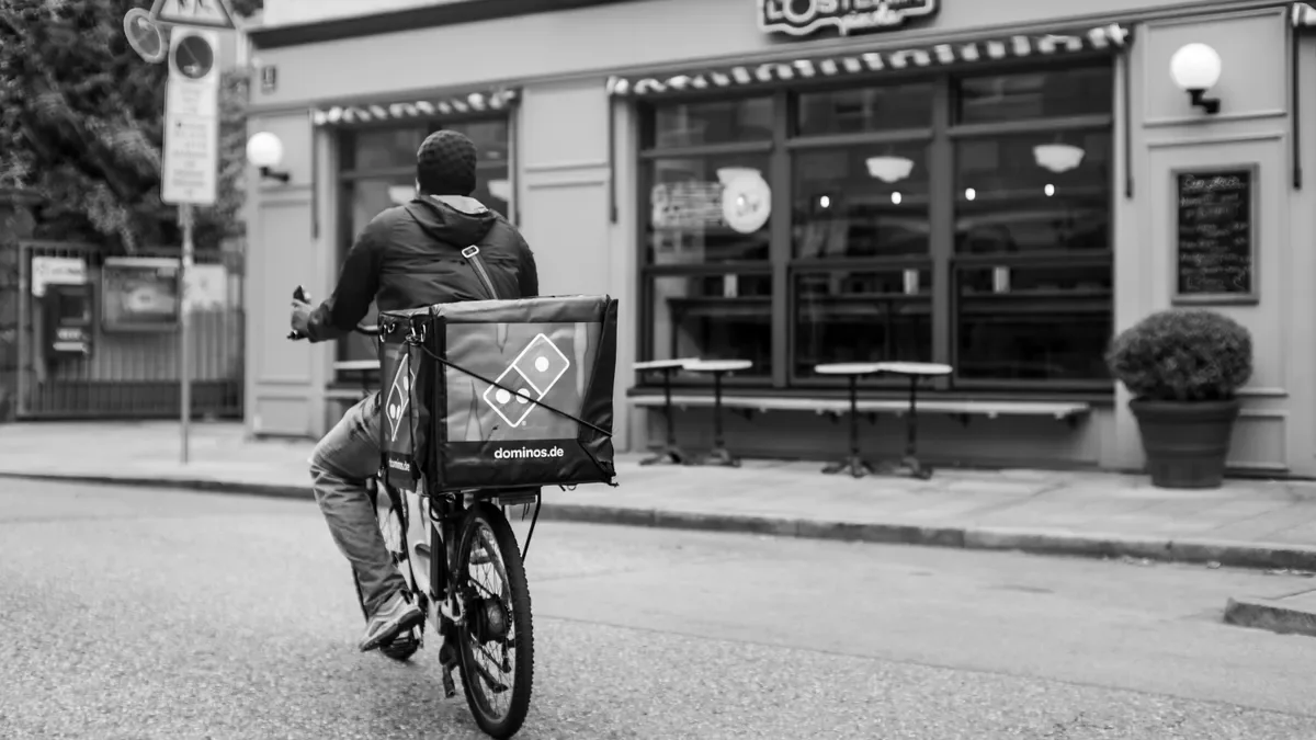Domino's delivery cyclist with a branded box on the back of his bike riding past a restaurant