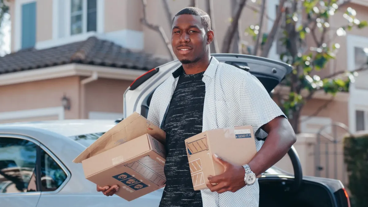 Delivery driver carrying two parcels from the open boot of a silver car parked outside a house