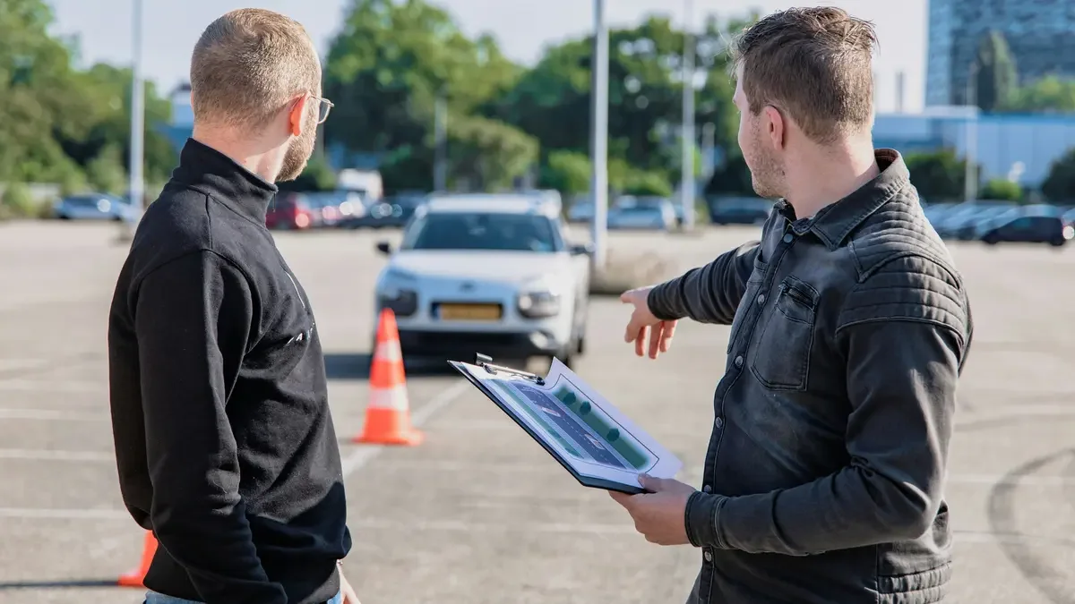 Driving instructor holding a clipboard and pointing towards a car and traffic cones during an assessment
