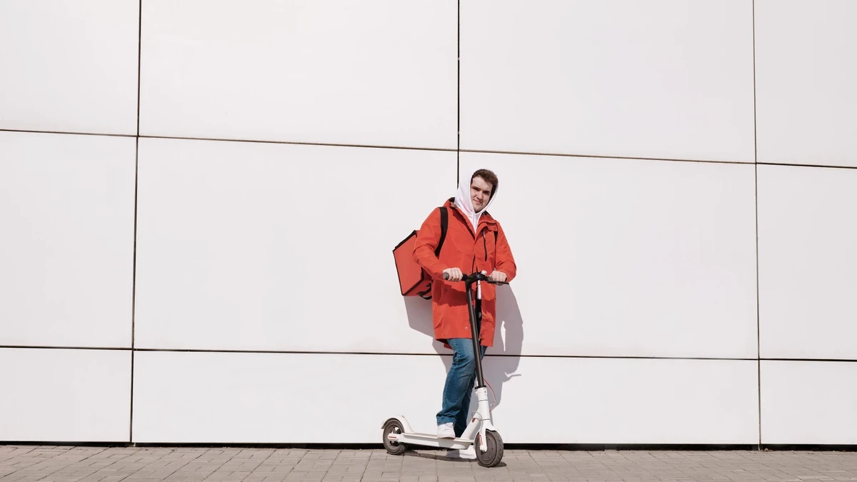 Food courier in a red jacket with a red thermal bag standing on an electric scooter by a white wall