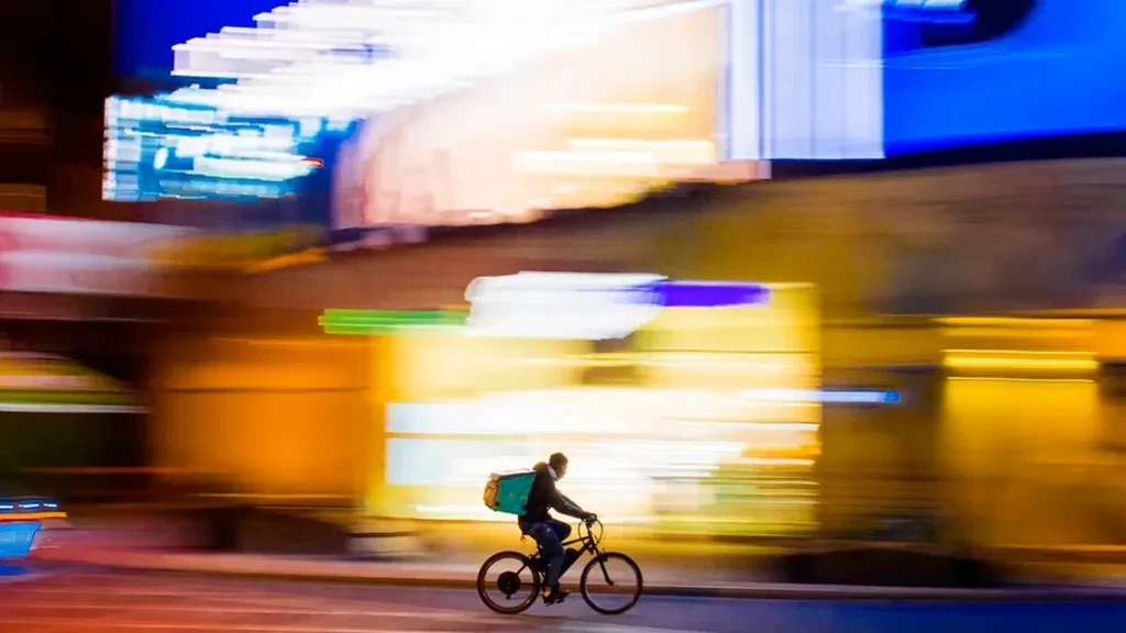 Food delivery cyclist riding past blurred, brightly lit shopfronts at night