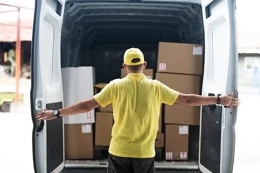 Courier in a yellow polo shirt and cap opening the rear doors of a van loaded with cardboard parcels