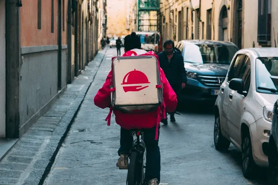 Food delivery cyclist with a red thermal backpack riding down a narrow street