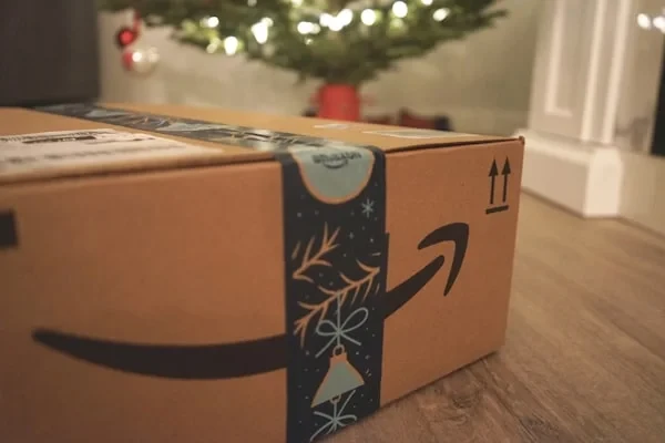 An Amazon parcel sealed with festive tape on a wooden floor, with a decorated Christmas tree out of focus behind it