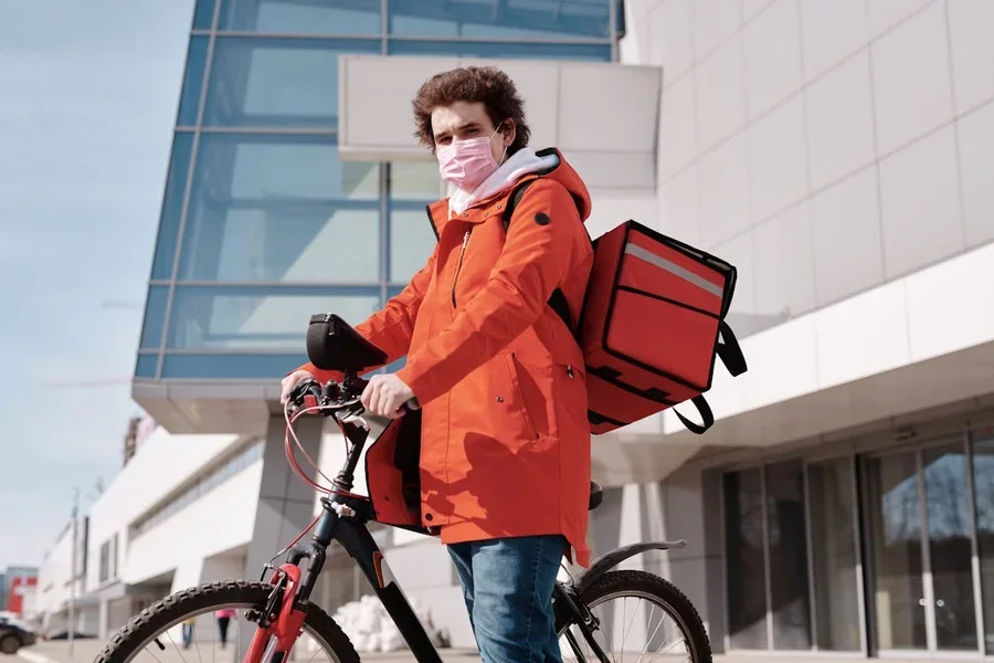 Courier with a red delivery backpack standing beside a bicycle outside an office building