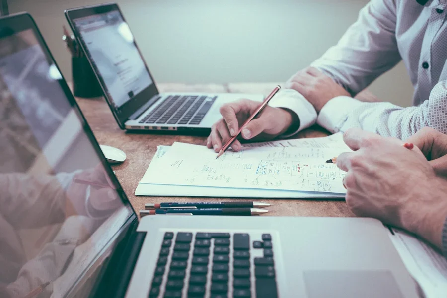 Two people reviewing insurance paperwork with a pen between two laptops on a desk