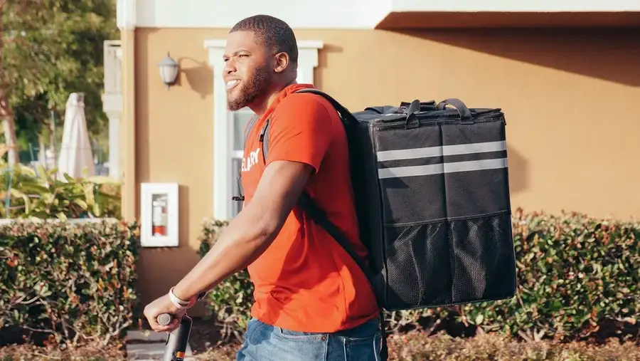Delivery rider with a large thermal backpack standing over an electric scooter outside a house