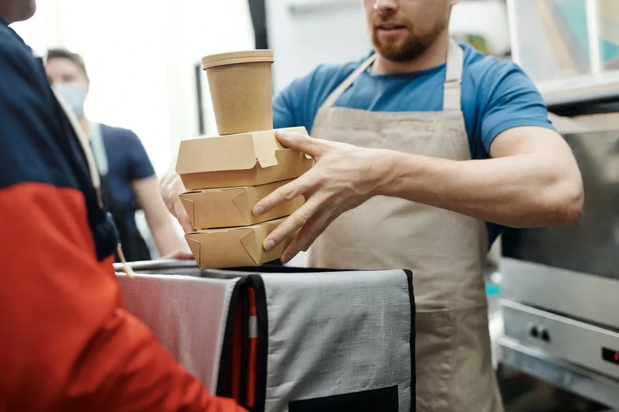 Kitchen worker in an apron stacking takeaway boxes and a paper cup on top of a courier's thermal bag