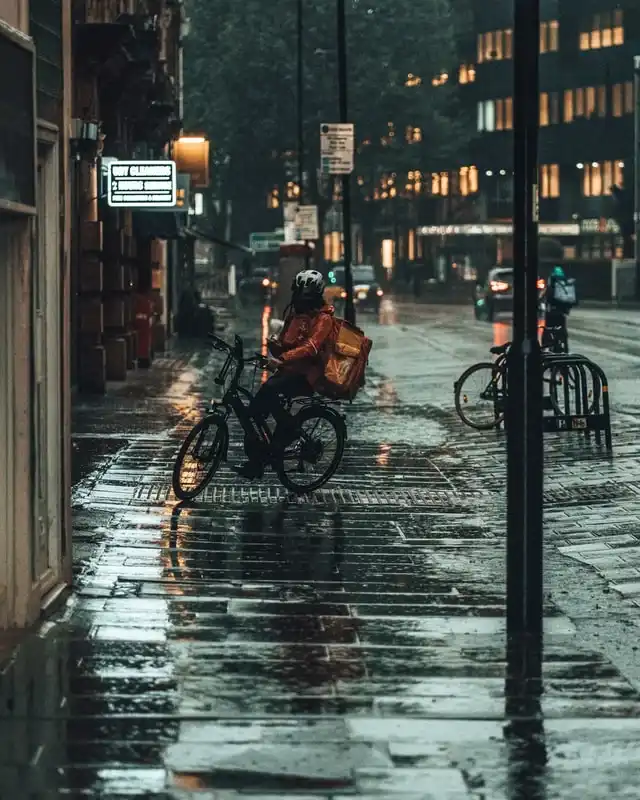 Delivery cyclist with a thermal bag riding across wet paving on a rainy city street