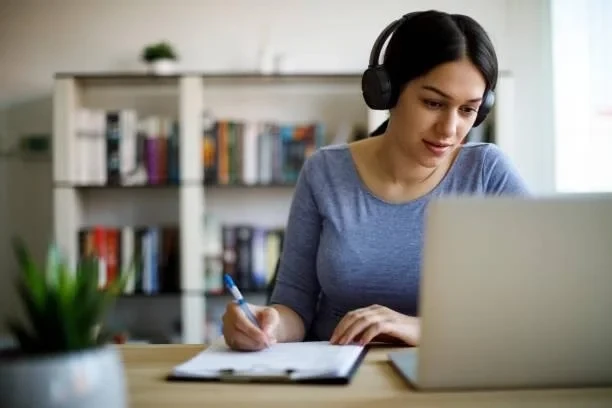 Woman studying at a laptop with headphones on, taking notes