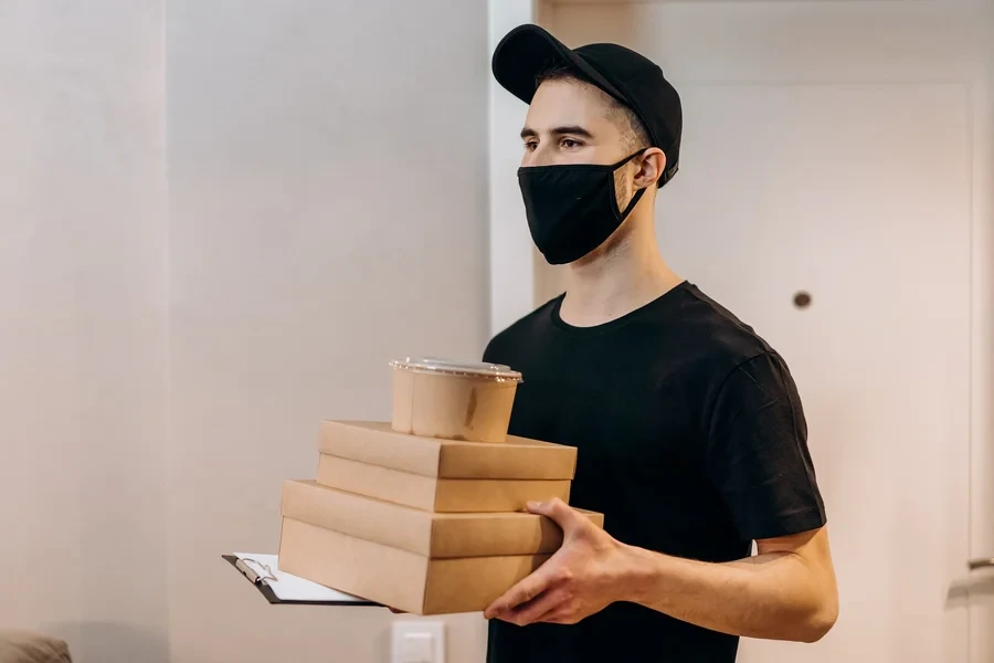Masked food courier in a black cap carrying stacked takeaway boxes on a clipboard