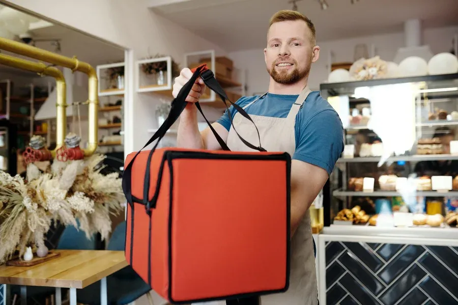 Smiling courier holding up a red thermal delivery bag inside a bakery