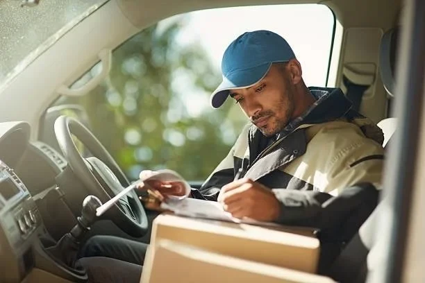 Courier sitting in the driver's seat of a van, signing a delivery note with a parcel on his lap