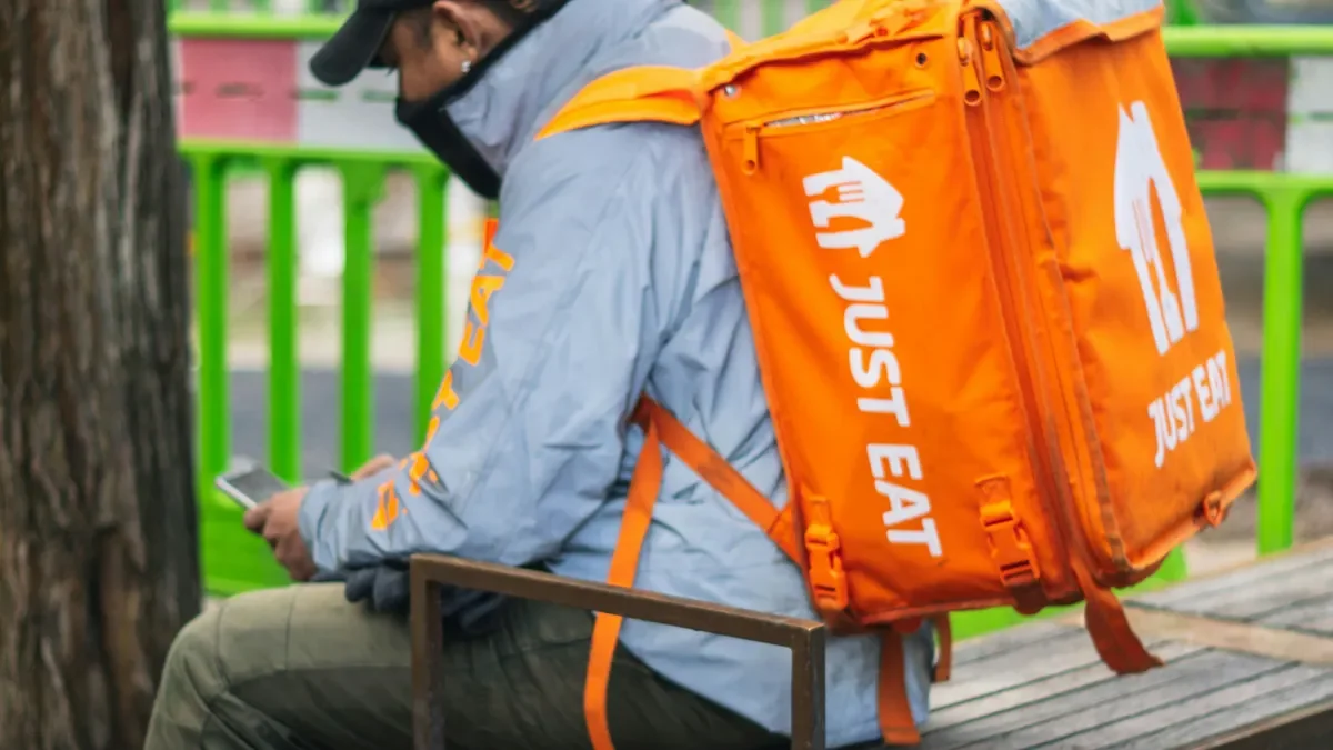 Just Eat courier sitting on a bench checking his phone, orange delivery backpack over his shoulders