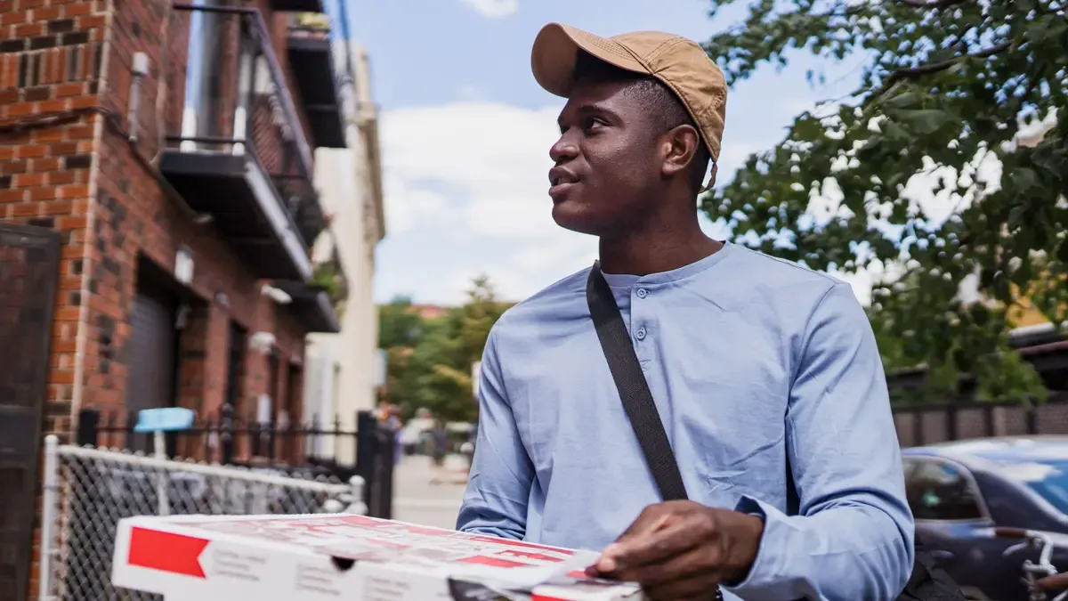 Pizza courier in a cap carrying a boxed pizza along a residential street on a sunny day