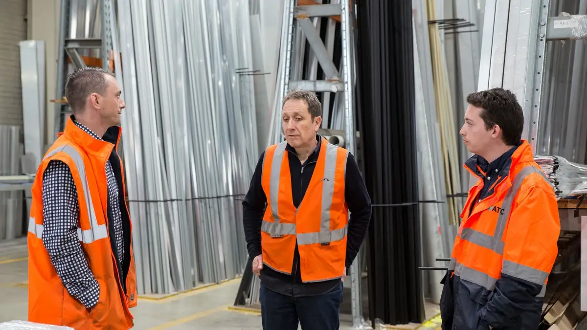 Three colleagues in orange hi-vis vests talking in a warehouse aisle lined with racks of metal lengths