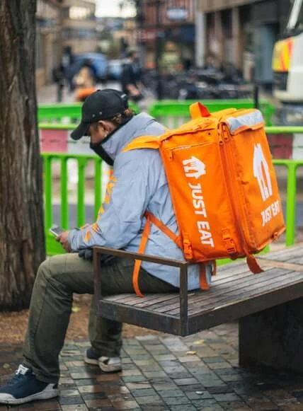 Just Eat courier sitting on a bench with a large orange insulated delivery backpack, checking his phone