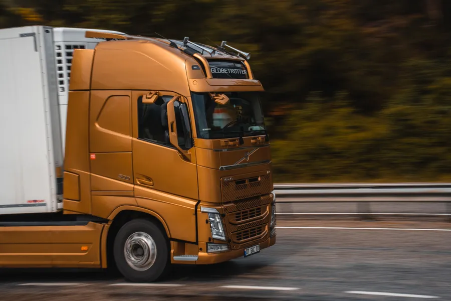 Copper-coloured Volvo FH articulated lorry pulling a white refrigerated trailer along a tree-lined main road