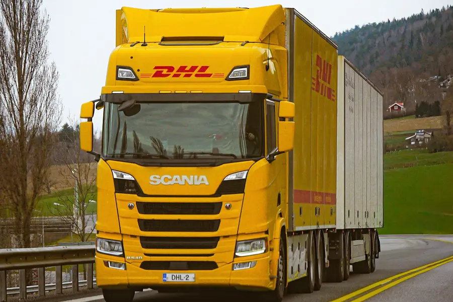 Yellow DHL Scania articulated lorry pulling curtainsided trailers along a rural main road past green hillsides