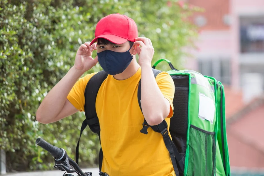 Courier in a red cap and face mask with a green insulated delivery backpack, standing with a bicycle
