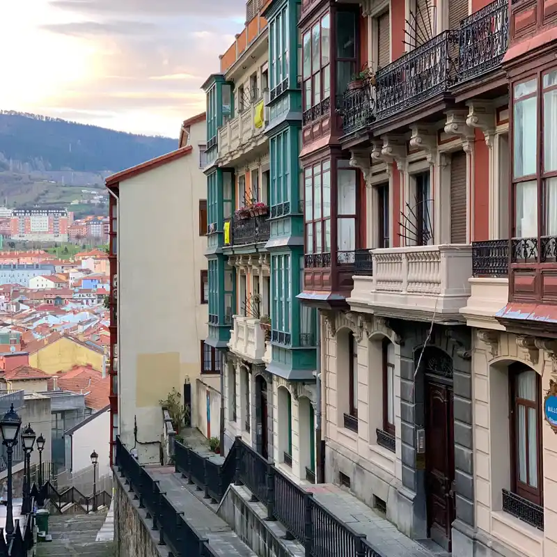Escalinata con farolas y barandillas de hierro entre edificios de miradores en Bilbao, con los tejados de la ciudad y los montes al fondo