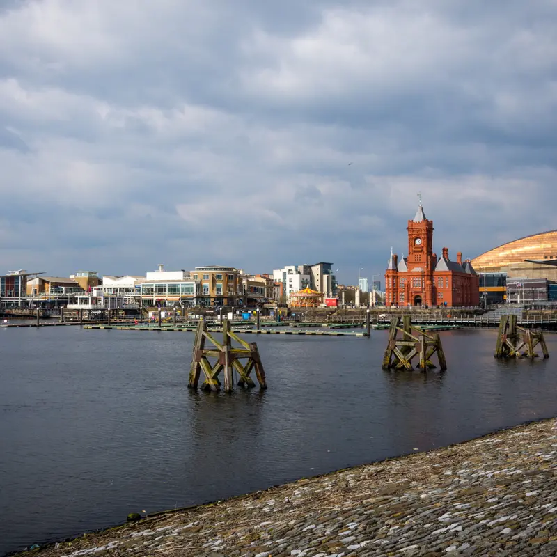 Cardiff Bay under a cloudy sky, with the red brick Pierhead Building, the Mermaid Quay waterfront and the curved roof of the Wales Millennium Centre across the water