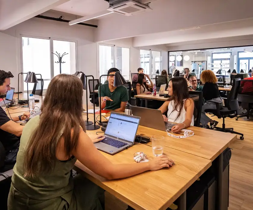Teammitglieder arbeiten mit Laptops an gemeinsamen Tischen im Büro von Service Club, im Hintergrund hohe Fenster