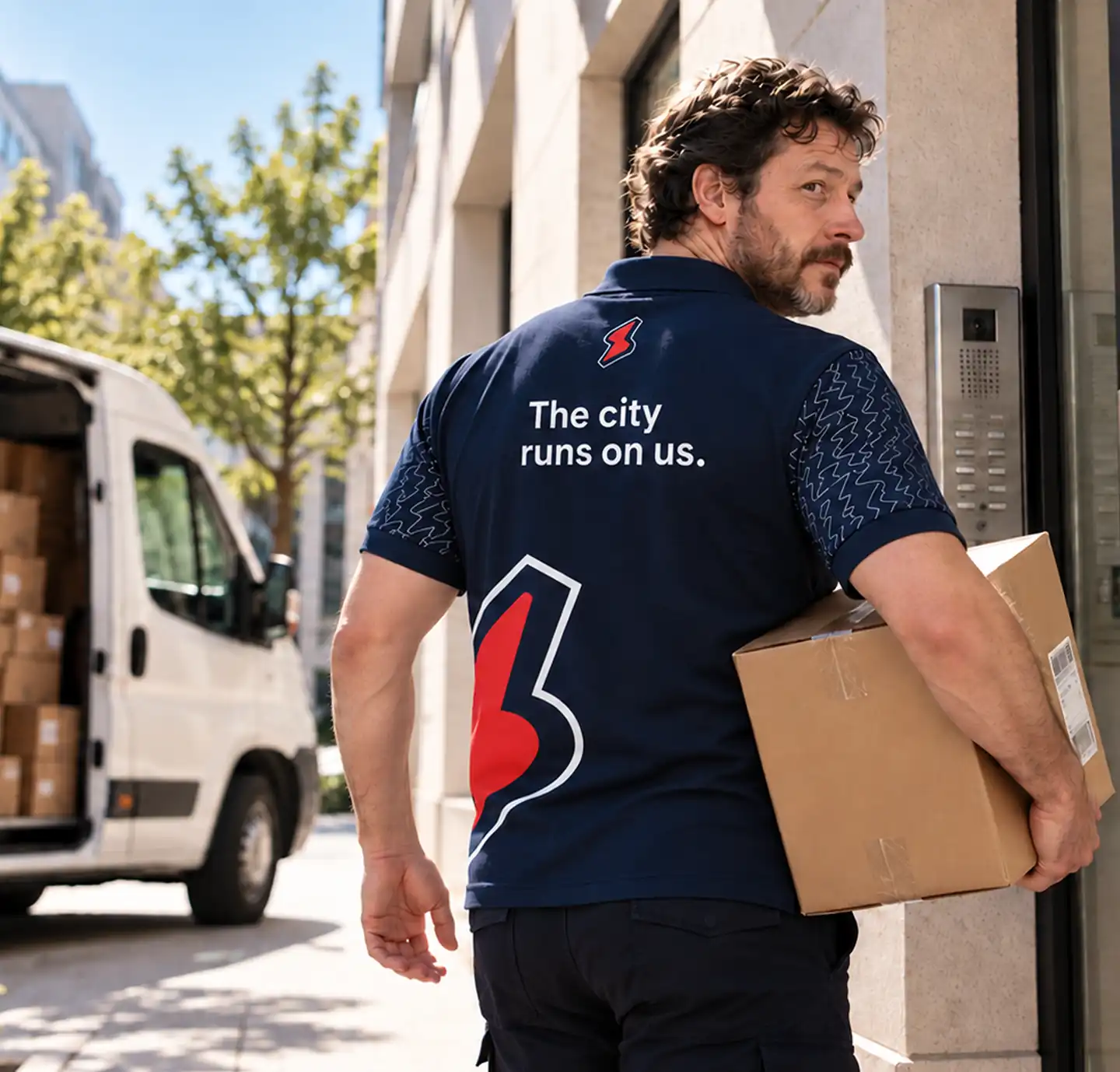 Courier in a Service Club polo with a parcel under his arm at a building entrance, an open van behind him