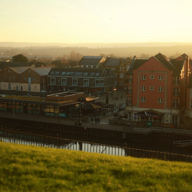 Cafes and bars on Exeter Quay at sunset, seen from the grassy bank across the water, with red brick flats beside them and hills on the horizon