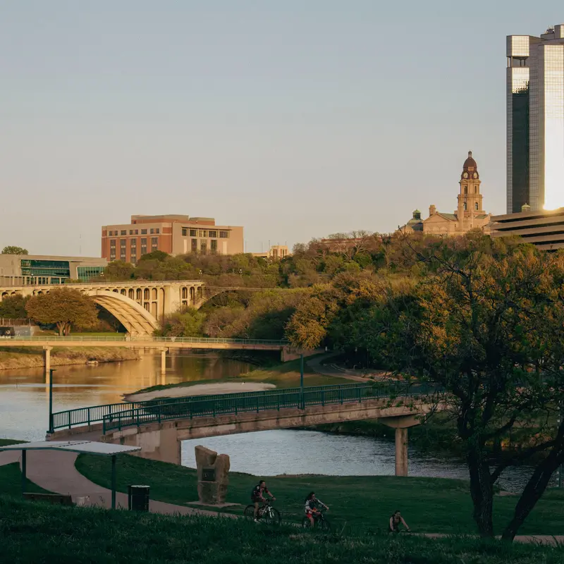 Downtown Fort Worth above the Trinity River at golden hour, with the Tarrant County Courthouse tower, an arched road bridge and cyclists on the riverside trail
