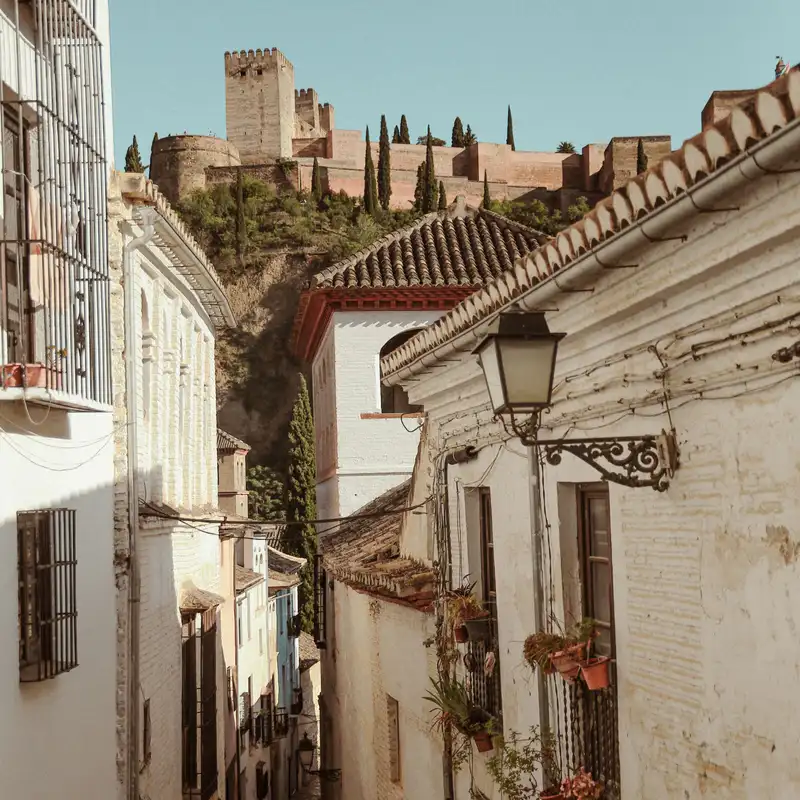 Callejón encalado del casco antiguo de Granada, con farol de forja y macetas en las ventanas, y la Alcazaba de la Alhambra entre cipreses en lo alto de la colina