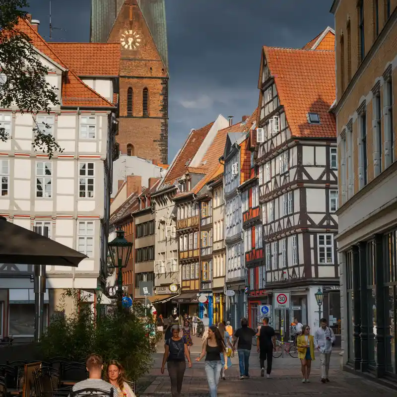 Fachwerkhäuser in der Altstadt von Hannover, Menschen spazieren durch die Fußgängerzone, dahinter der Backsteinturm der Marktkirche mit Uhr