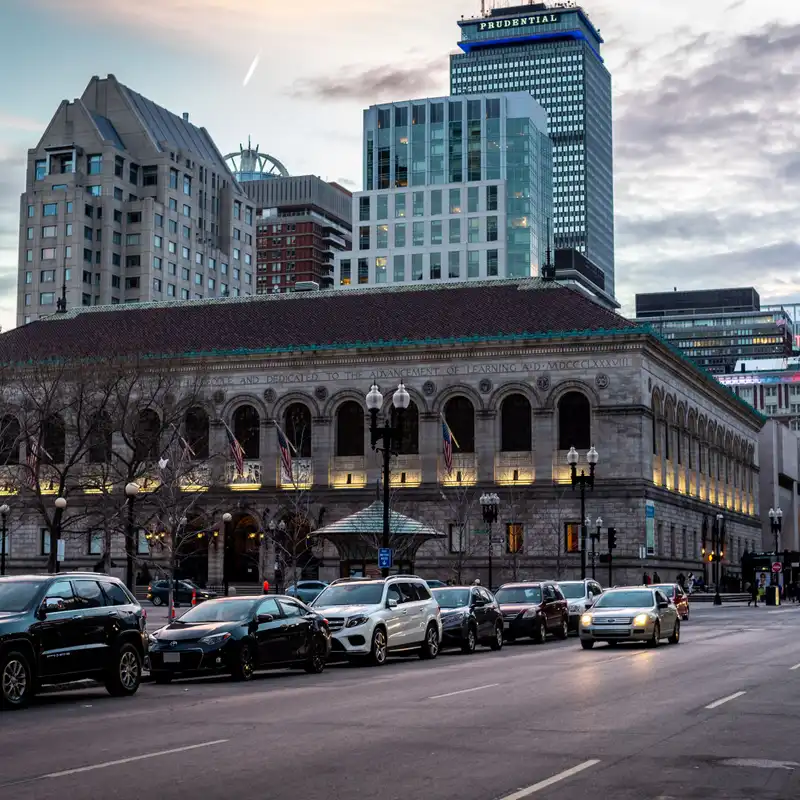Cars parked and driving past the Boston Public Library on the edge of Copley Square at dusk, with the Prudential Tower rising behind it