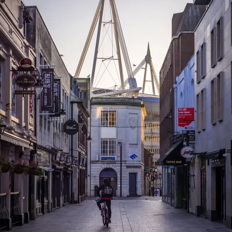 A cyclist rides along a paved lane in central Cardiff, past pubs and a tapas bar, with the masts of the Principality Stadium rising behind