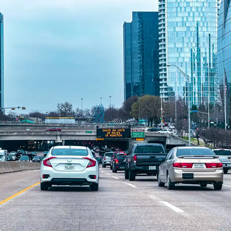 Traffic on a downtown Dallas freeway approaching the Pearl Street exit, with glass towers by the Arts District