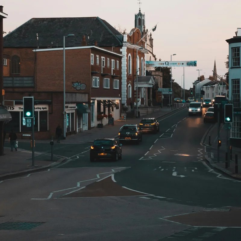 Cars with their headlights on driving up a street in central Exeter at dusk, under a banner pointing the way to the Quay, past red brick buildings and a white fronted building with a small tower