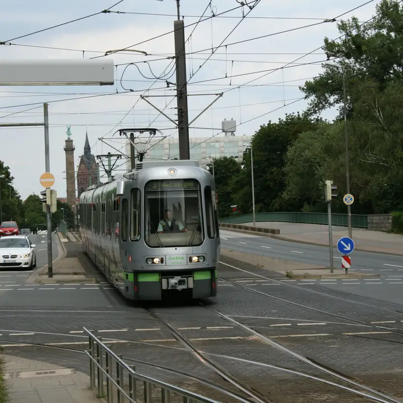 Eine silbergrüne Stadtbahn der Üstra fährt über die gepflasterte Gleistrasse, daneben Autos auf der Straße und im Hintergrund eine Säule mit Statue und ein Kirchturm