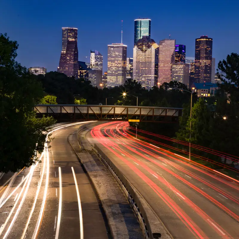 Evening traffic leaving red and white light trails on a parkway under a footbridge, with the downtown Houston skyline lit up behind