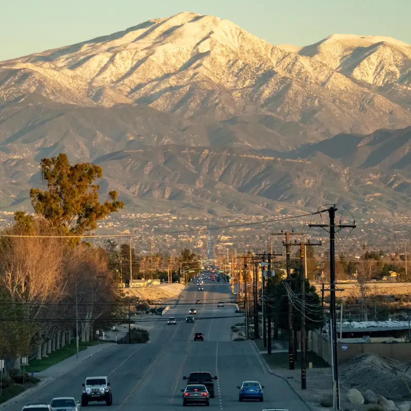 A wide road in the San Bernardino area at golden hour, traffic heading toward snow-capped mountains above the valley