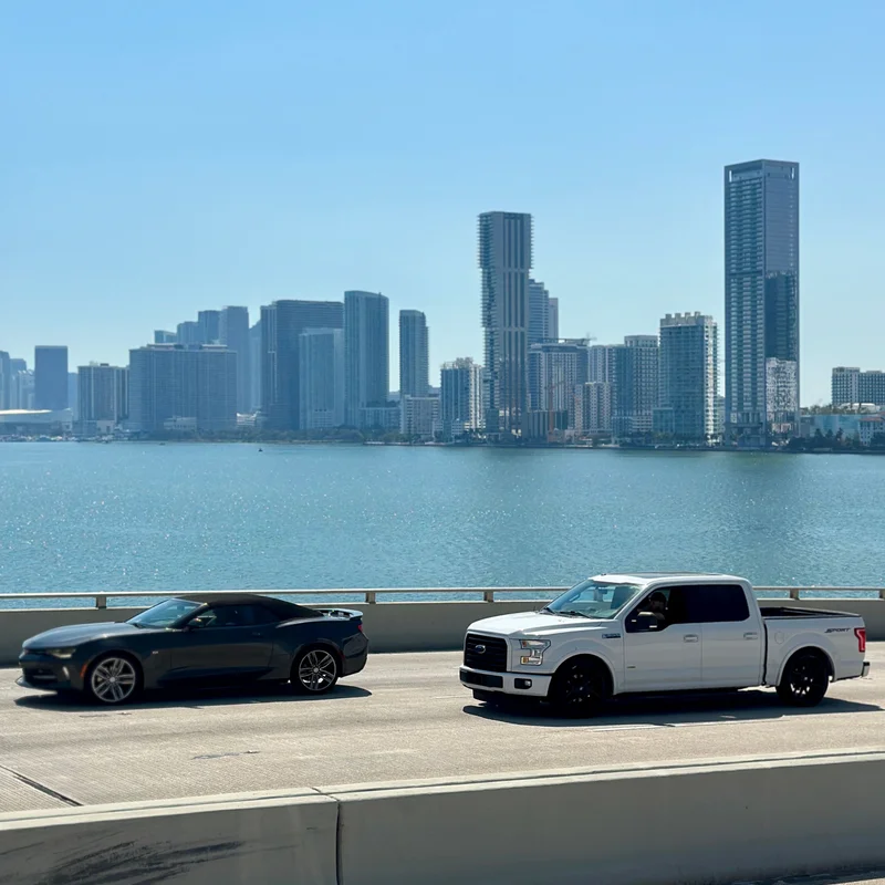 A car and a pickup truck crossing a causeway over Biscayne Bay, with the Miami skyline across the water