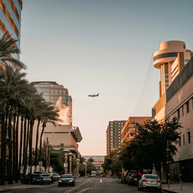 Palm trees line a downtown Phoenix street at dusk as cars drive toward the camera and a passenger jet descends between the office towers