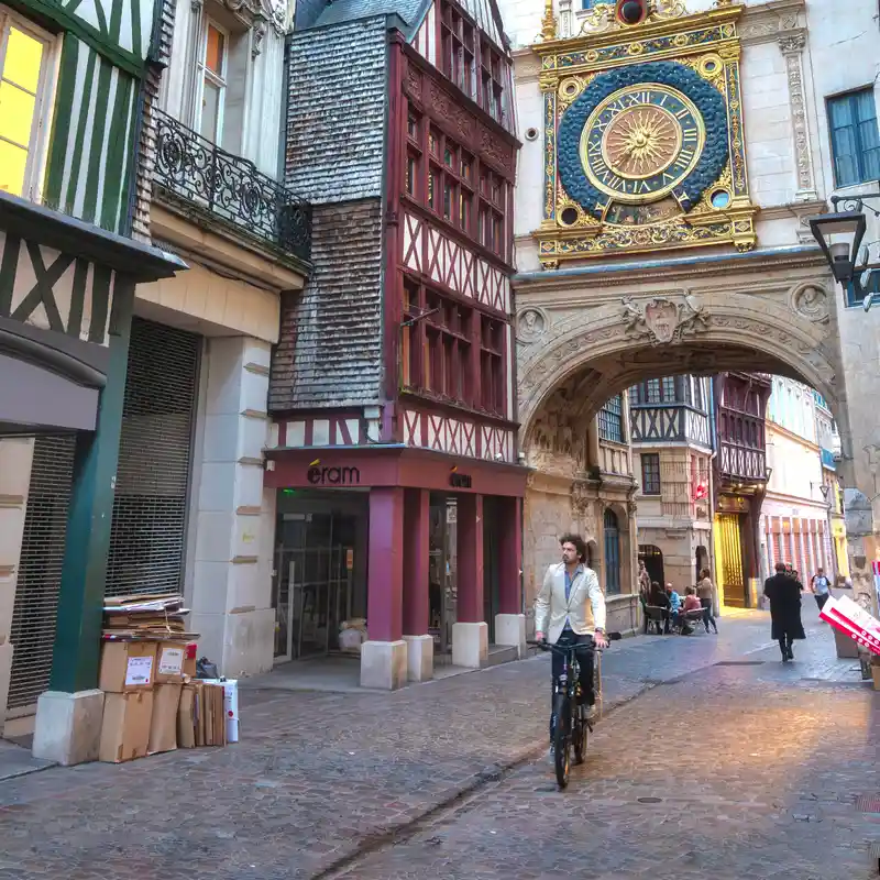 Une personne à vélo, en veste claire, descend la rue du Gros-Horloge, pavée et bordée de maisons à colombages, sous l'arche de l'horloge astronomique dorée